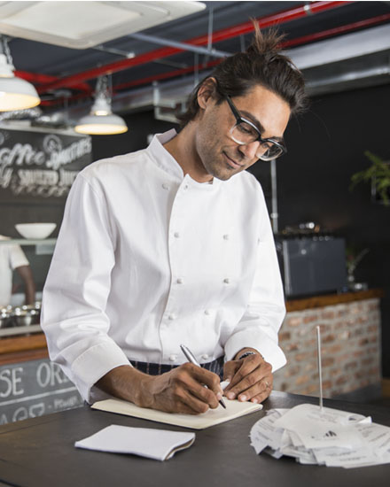 Un hombre con ropa blanca de chef haciendo una lista mientras está de pie en una cocina comercial.