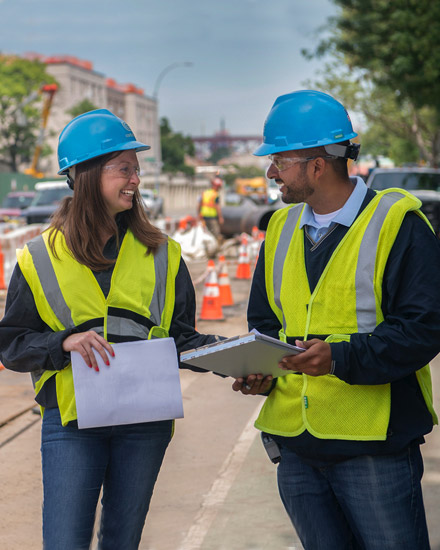 Two utility workers discuss plans on a city street.