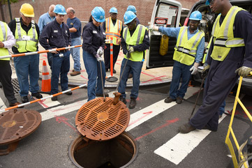 A crew of Con Edison workers are lifting a manhole cover. 
