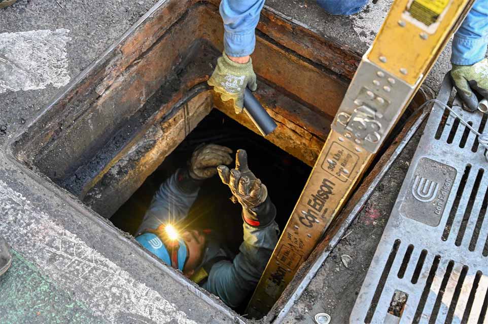 A Con Edison employee inside a maintenance hole reaches up for flashlight being handed to them.