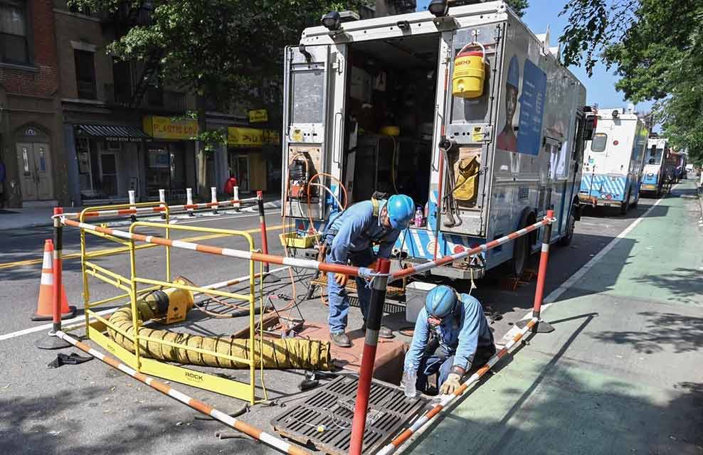 Two Con Edison workers remove a storm drain grate within a closed off service area on a city street.