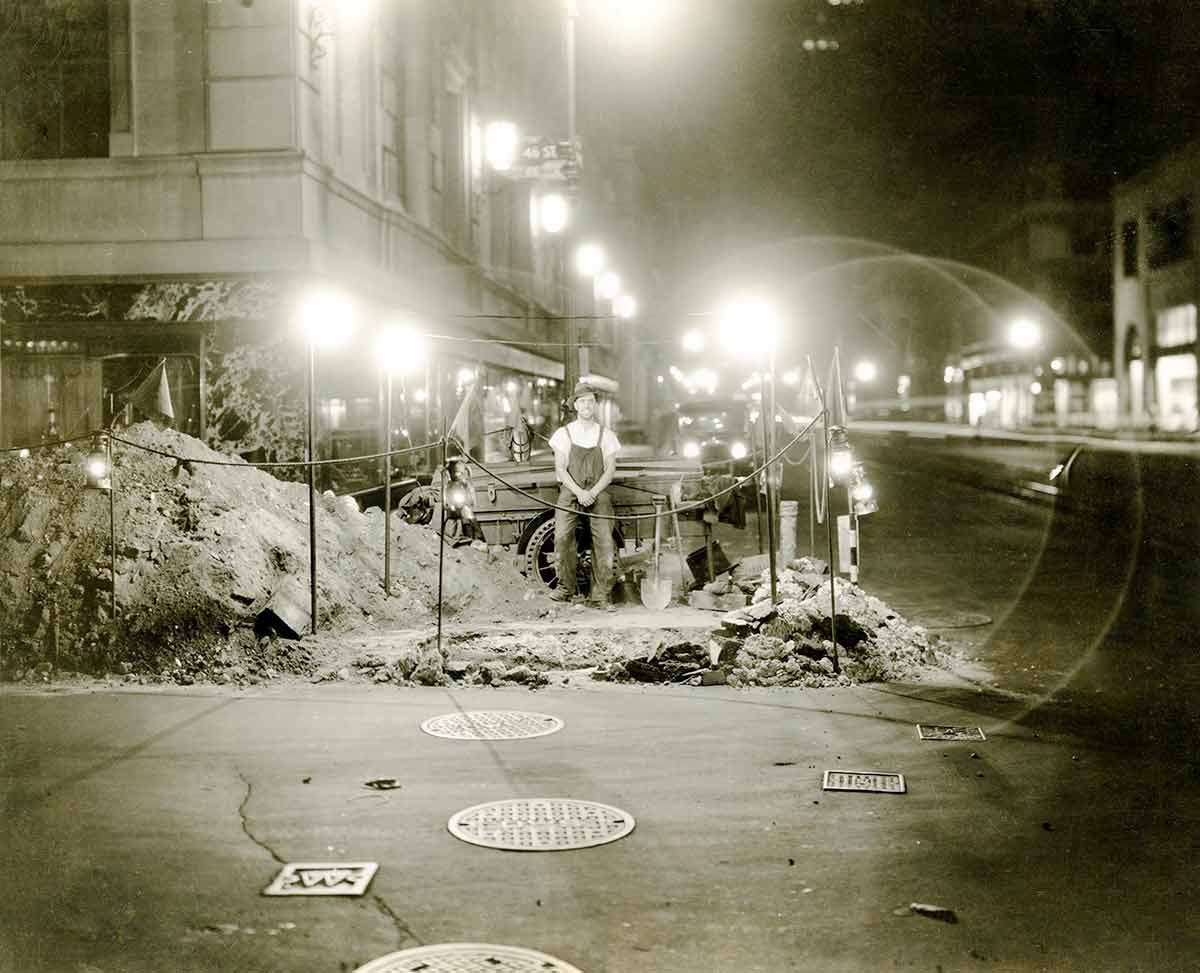 Fotografía en blanco y negro de un trabajador de Con Edison de pie frente a una excavación en una esquina de la ciudad de Nueva York.