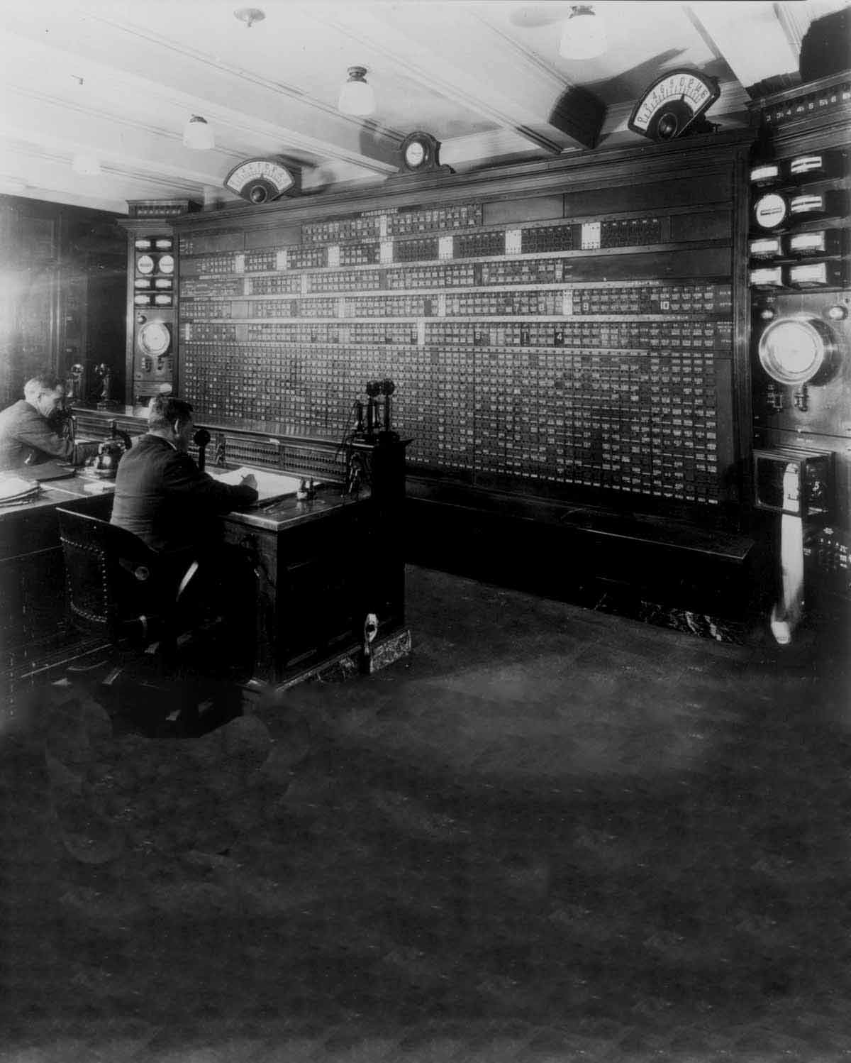 Fotografía en blanco y negro de dos trabajadores sentados observando una estación de monitoreo.