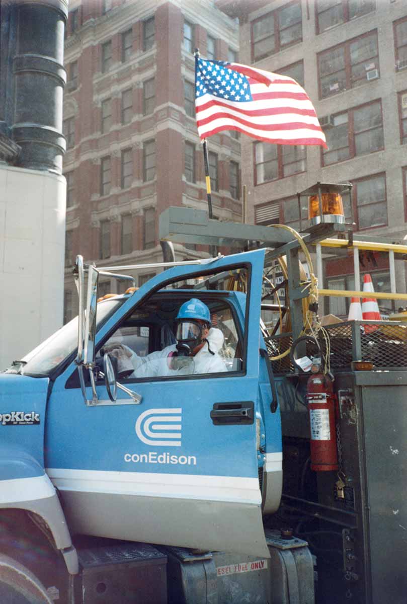 A Con Edison employee wearing a gas mask and hazmat suit sits in the driver seat of a work truck.