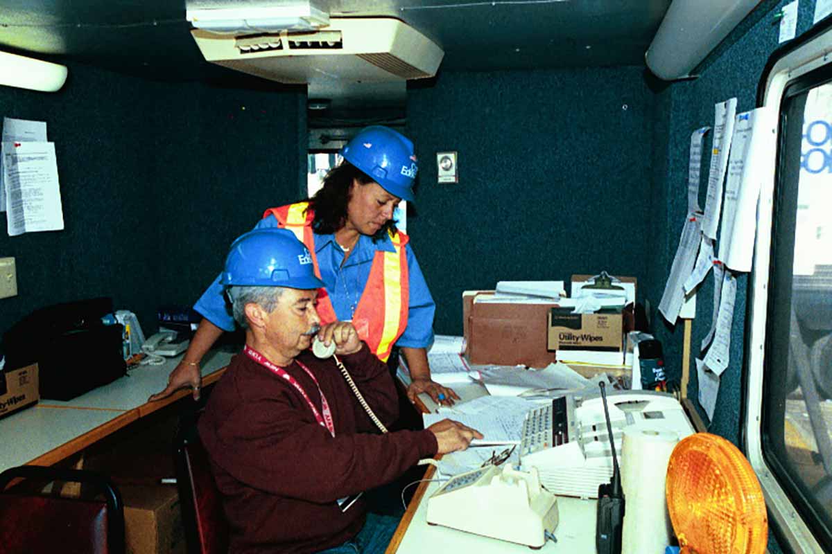 A Con Edison employee talks on the phone as another employee stands over them looking at diagrams on a desk.