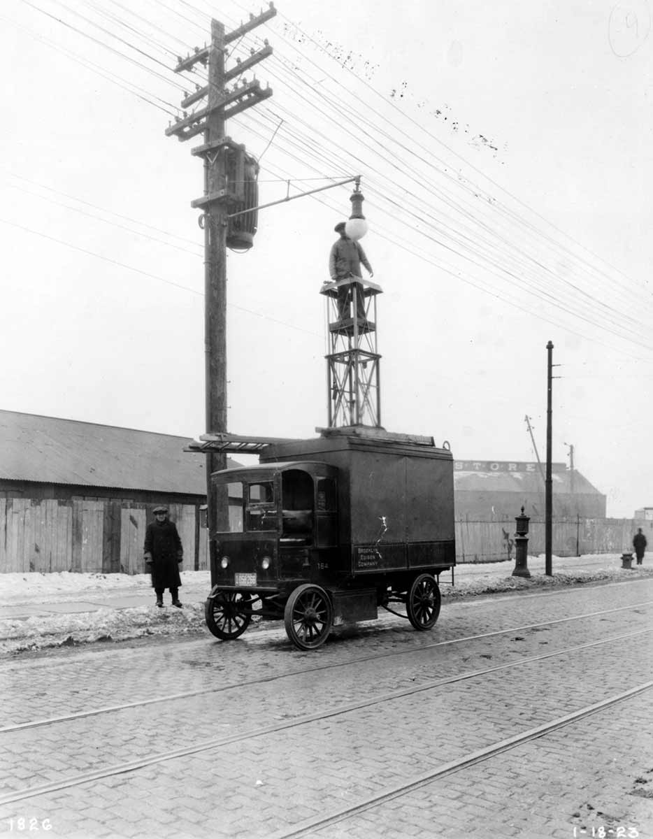 Black and white photo of a worker hoisted up a truck and platform to repair a streetlamp.
