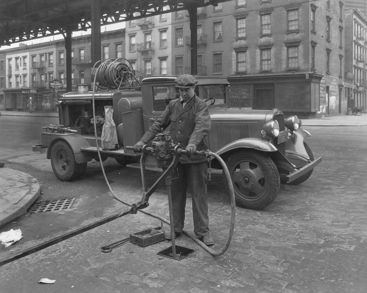 Black and white photo of a Con Edison worker connecting a hose to a pipe on a city street.