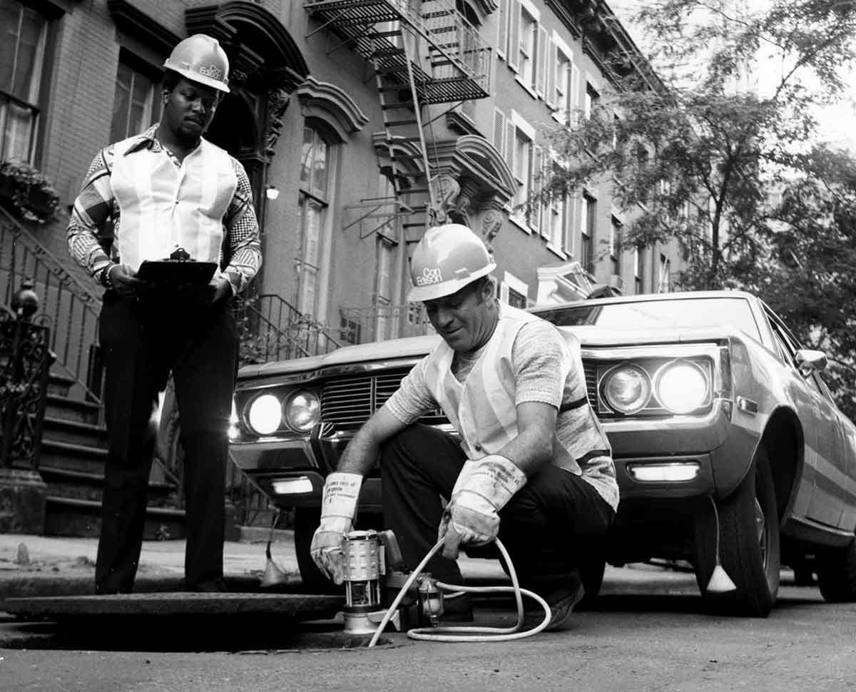 Black and white photo of two Con Edison workers taking measurements by an open manhole.