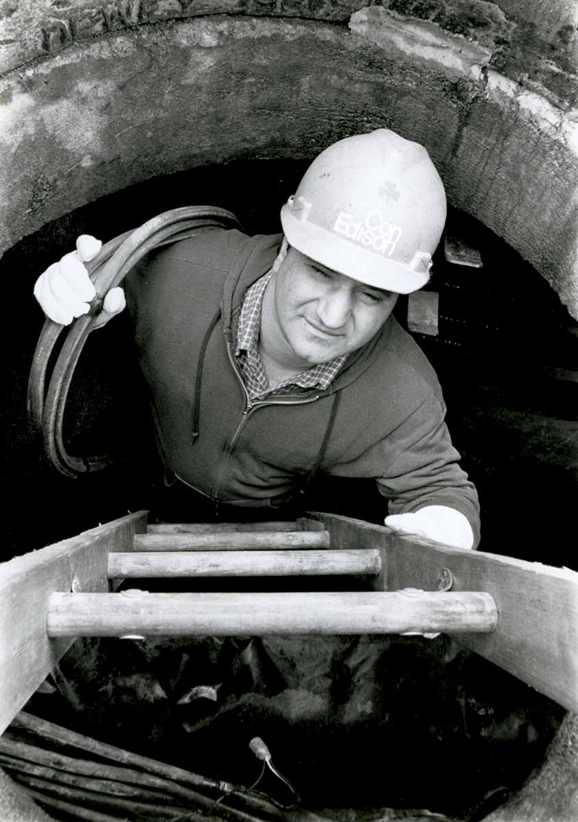 Black and white photo of a Con Edison worker climbing up a manhole while holding cable.
