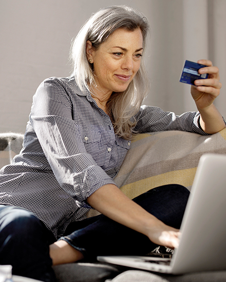A woman is sitting on a couch and using her credit card to pay a bill on her laptop.