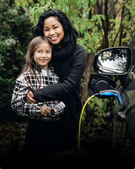 A woman embraces her young daughter, while her electric vehicles is being charged. 