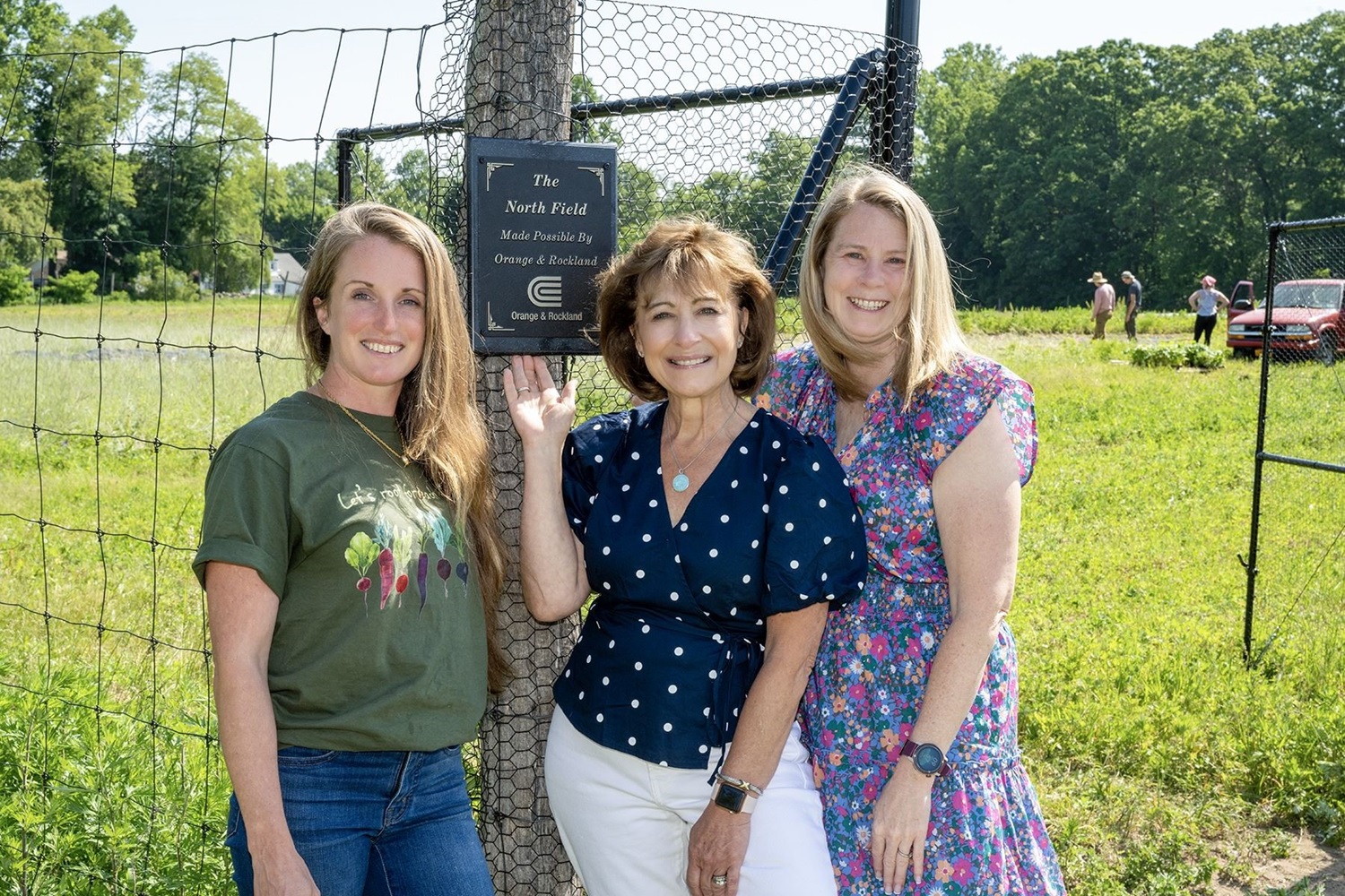 Three women stand on field next to plaque honoring O&R. 