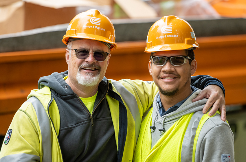 Two Orange & Rockland employees wearing safety gear pose with their arms over each other's shoulders.