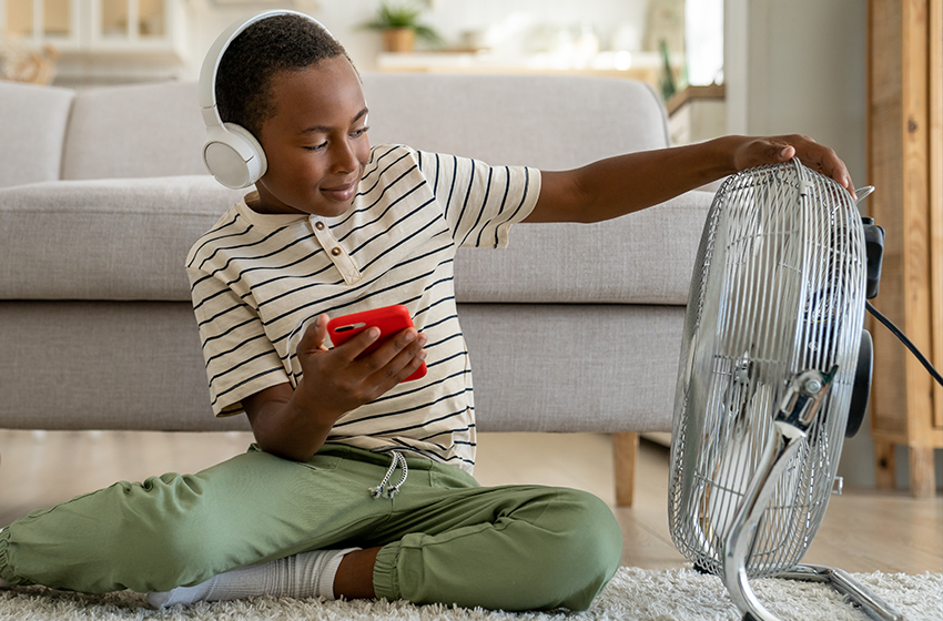 A child sits on the living room floor in front of a fan while listening to music on a smartphone.